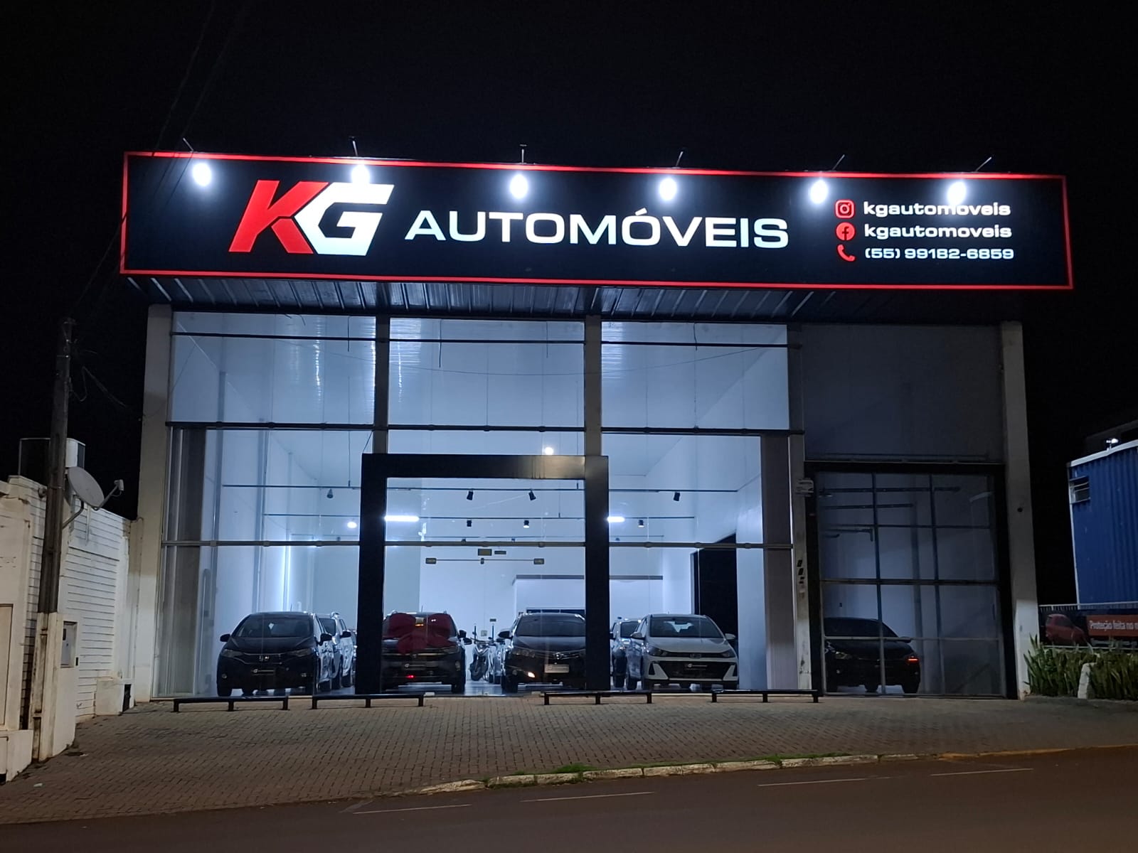 Fachada da KG Automóveis à noite, com o letreiro iluminado e os veículos dentro da loja